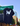 A black short-sleeve T-shirt is shown hanging outdoors against a city street background with a green Madison Street sign and buildings. The shirt has a large white dripping letter W graphic on the back with smaller white text underneath reading 'WORN BY THE CHOSEN'.